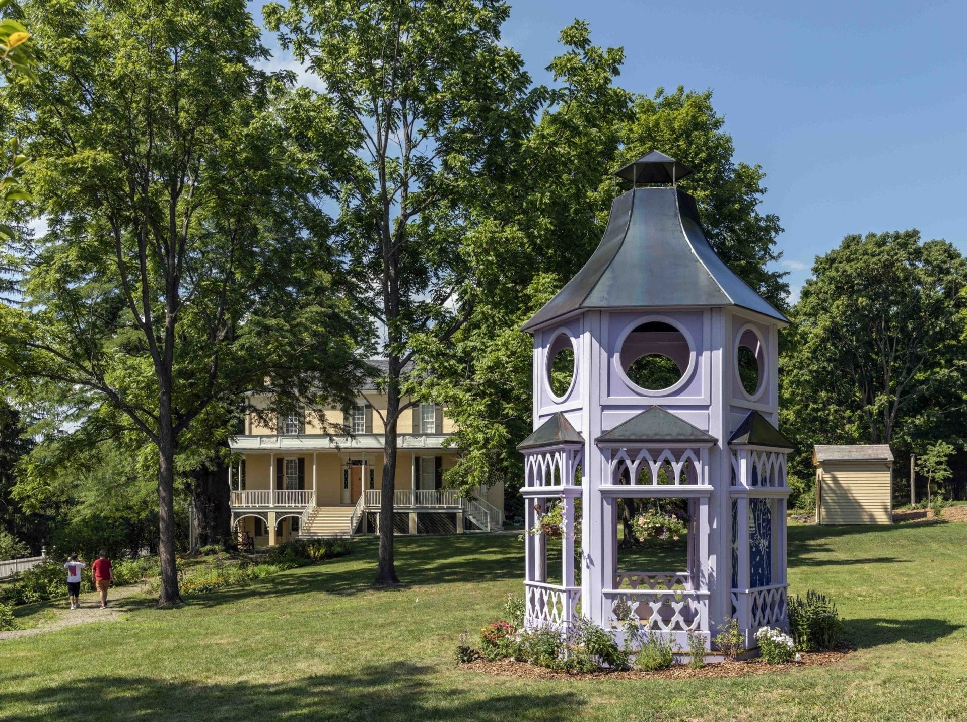 A purple gazebo stands in a sunny park surrounded by lush green trees, reminiscent of the enchanting New York sculpture parks worth visiting. In the background, a large yellow house with a porch is visible. Two people stroll peacefully along a path on the left. A purple gazebo stands in a sunny park surrounded by lush green trees, reminiscent of the enchanting New York sculpture parks worth visiting. In the background, a large yellow house with a porch is visible. Two people stroll peacefully along a path on the left.