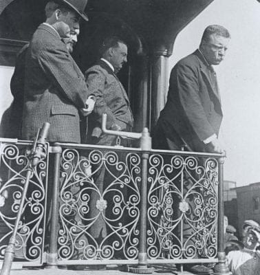 A historical black-and-white photo captures a man in a suit and hat standing on a decorative wrought iron balcony, exuding a presidential air. Another leans on the railing, addressing the crowd gathered below. The scene suggests an early 20th-century event filled with anticipation and public discourse. A historical black-and-white photo captures a man in a suit and hat standing on a decorative wrought iron balcony, exuding a presidential air. Another leans on the railing, addressing the crowd gathered below. The scene suggests an early 20th-century event filled with anticipation and public discourse.