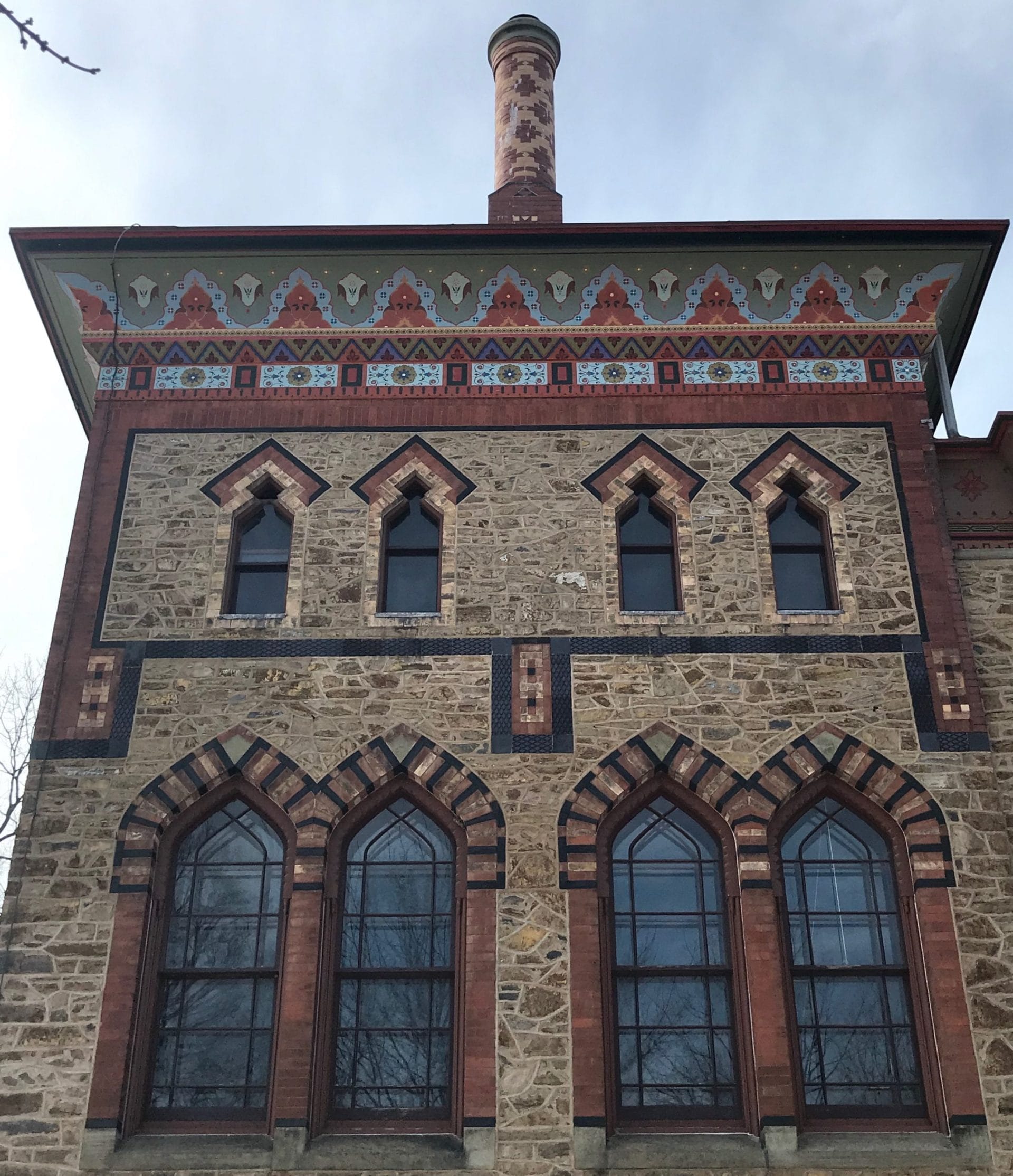 The image captures a historic building facade reminiscent of visiting Hudsons Olana State Historic Site, with intricate stonework. It features symmetrical arched windows and colorful decorative patterns beneath the roof, topped by a tall, patterned chimney against a cloudy sky. The image captures a historic building facade reminiscent of visiting Hudsons Olana State Historic Site, with intricate stonework. It features symmetrical arched windows and colorful decorative patterns beneath the roof, topped by a tall, patterned chimney against a cloudy sky.