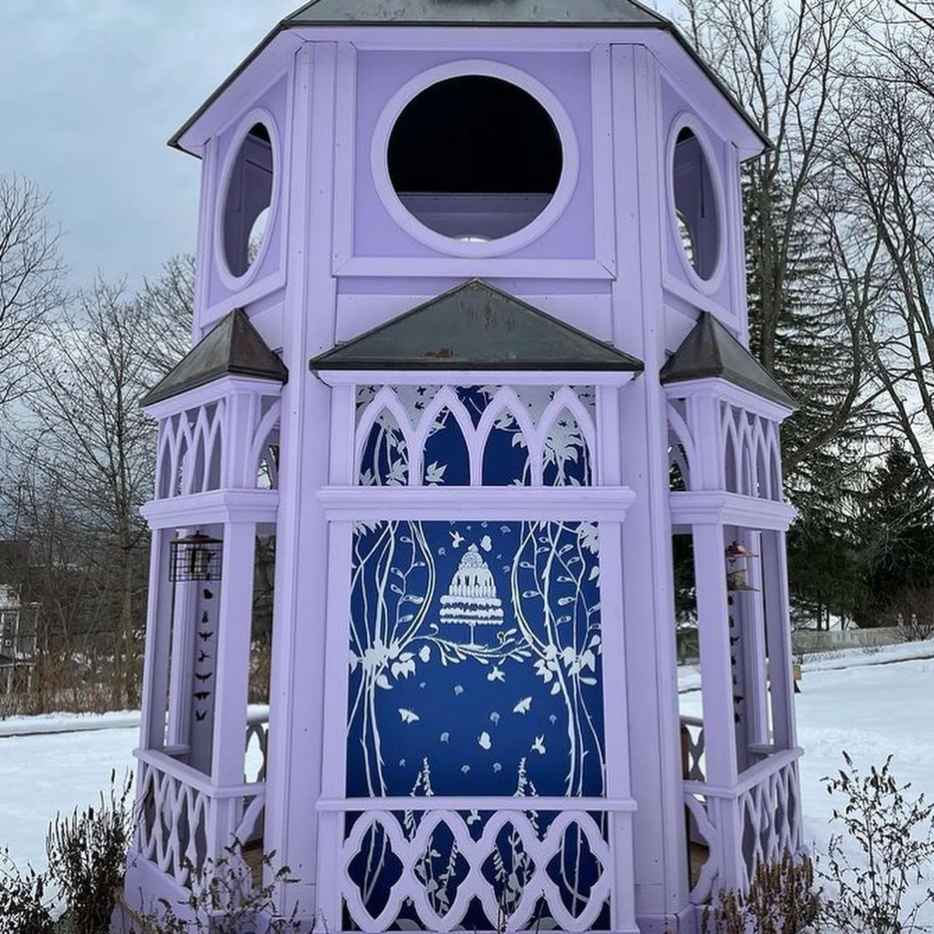 A tall, lavender-colored gazebo stands in a snowy landscape, resembling a pollinator pavilion with its intricate cut-out designs. Trees and birds embellish the blue centerpiece, while leafless trees and a cloudy sky form the serene backdrop. A tall, lavender-colored gazebo stands in a snowy landscape, resembling a pollinator pavilion with its intricate cut-out designs. Trees and birds embellish the blue centerpiece, while leafless trees and a cloudy sky form the serene backdrop.