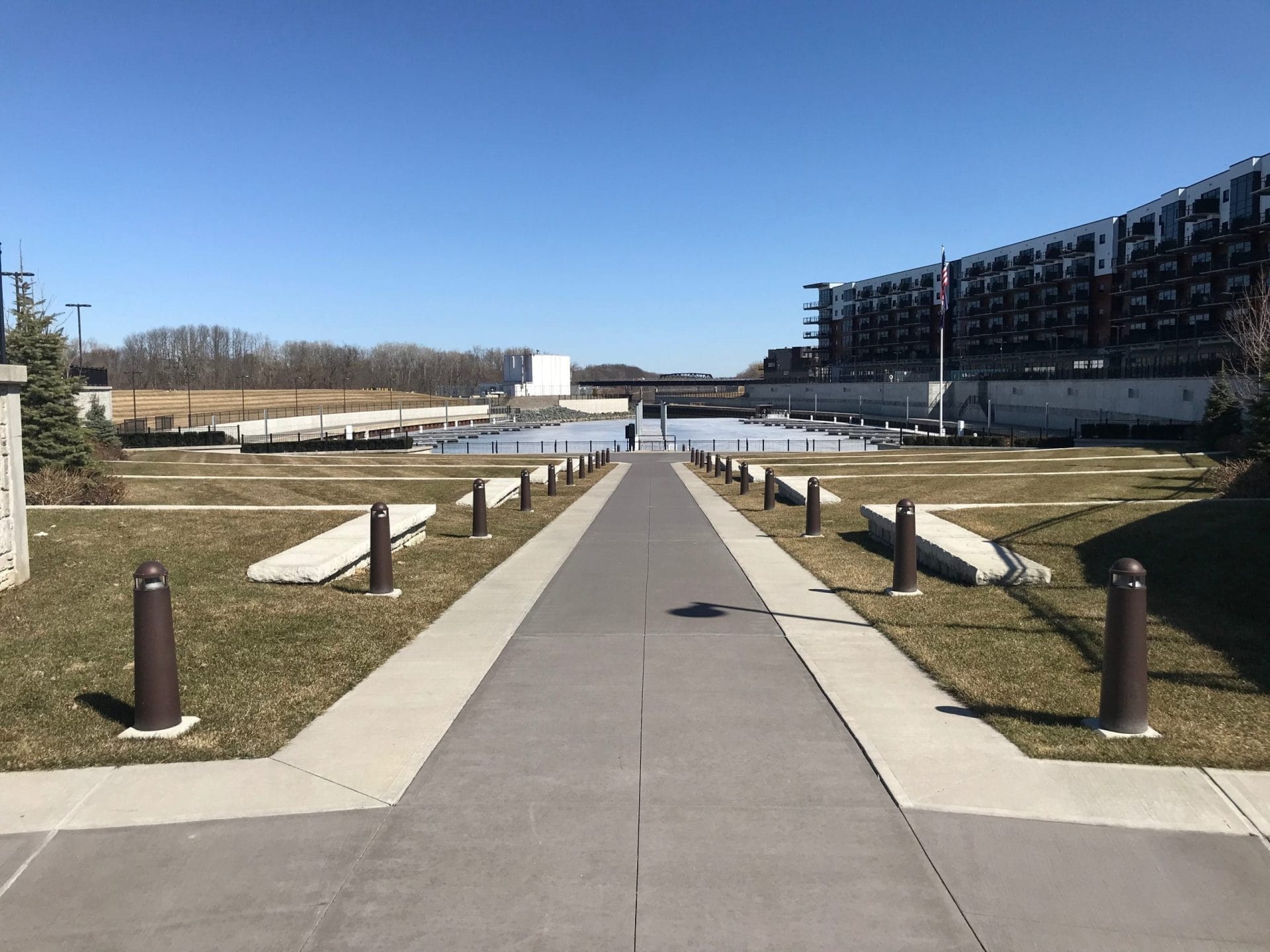 A paved pathway leads to a waterfront with the modern Rivers Casino & Resort on the right side. The scene includes grass on both sides of the path, bollards along the edges, and a clear blue sky.