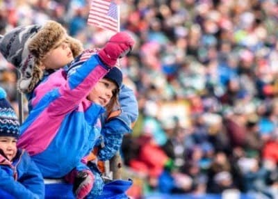 Children in colorful winter clothing enthusiastically watch an event, with one child waving a small American flag. The vibrant scene captures the spirit of a winter getaway to Vermont, made lively by the blurred crowds in the background.