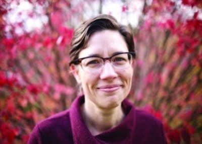 Smiling in front of vibrant red and pink foliage, a person with short hair and glasses wears a maroon top, evoking the serene beauty of a Vermont getaway.