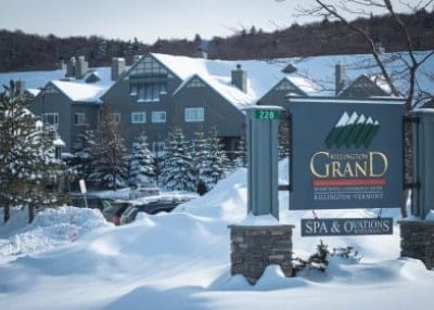 A large building is surrounded by snow-covered trees and mountains. In the foreground, a sign reads Killington Grand Resort Hotel Spa & Oneations. Snow blankets the ground and rooftops, highlighting a winter setting—a perfect get away to Vermonts serene beauty.