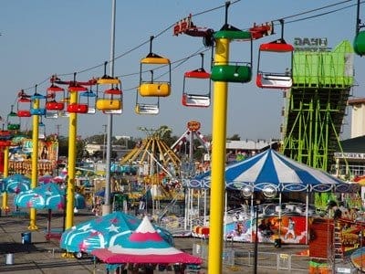 A vibrant amusement park reminiscent of the New York State Fair, featuring colorful cable cars on overhead tracks, a striped carousel, and various rides in the background. Bright tents and attractions fill the scene, capturing the lively, festive atmosphere. A vibrant amusement park reminiscent of the New York State Fair, featuring colorful cable cars on overhead tracks, a striped carousel, and various rides in the background. Bright tents and attractions fill the scene, capturing the lively, festive atmosphere.