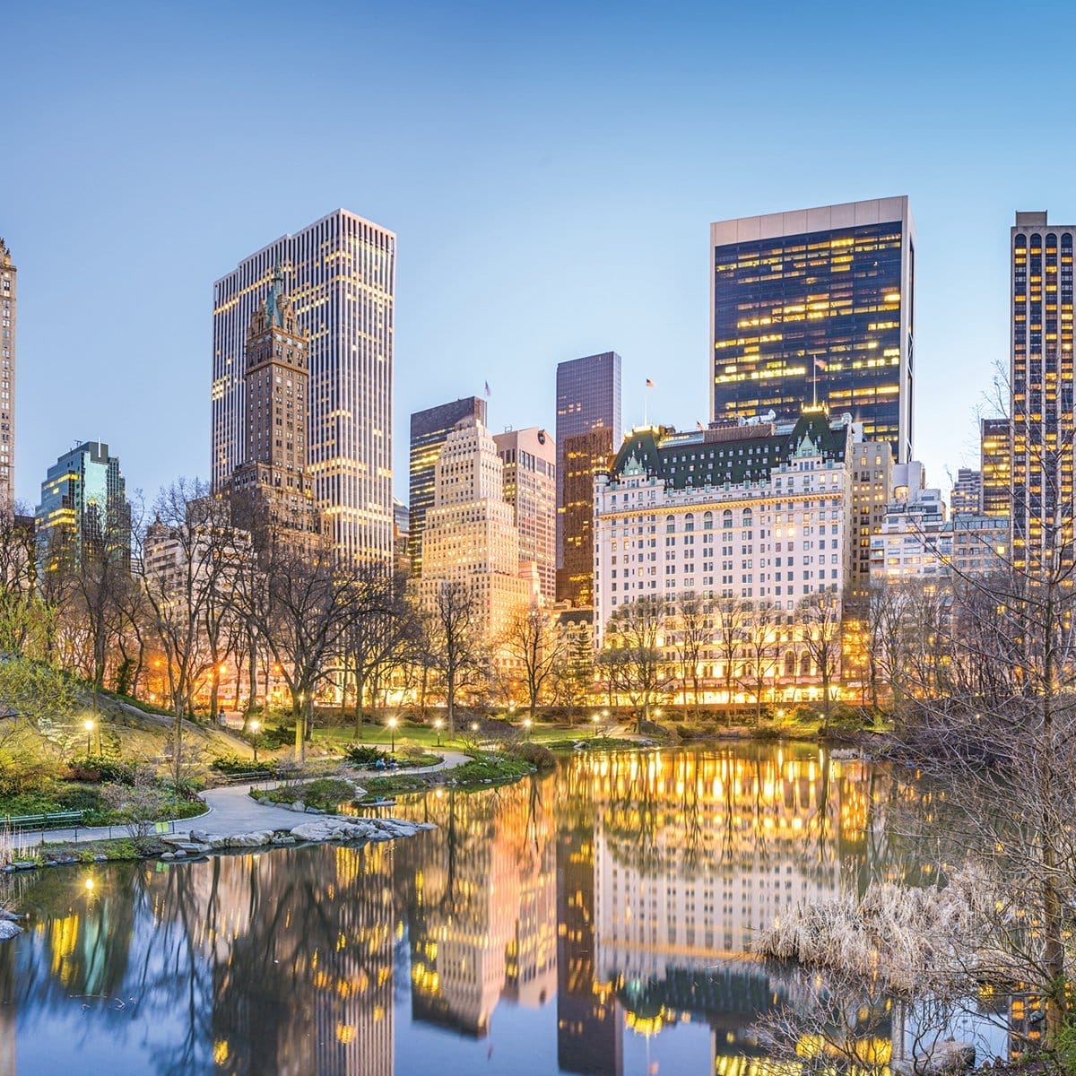 City skyline view from a park featuring tall, illuminated buildings reflecting in a calm body of water. Leafless trees are scattered in the foreground under a clear, twilight sky.