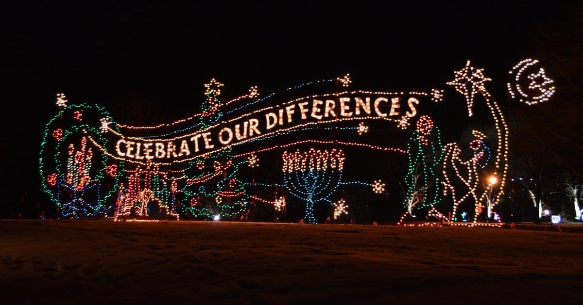 A vibrant holiday lights display at night showcases the message Celebrate Our Differences with festive symbols, including a Christmas tree, menorah, and crescent moon with star.