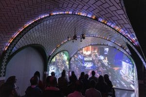 A group of people sit under an arched, tiled ceiling at RiseNY, watching a large, curved screen displaying a nighttime cityscape with bright lights and busy streets.