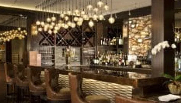 A modern bar in Saratoga Springs features stylish pendant lights hanging above a sleek countertop. Leather stools line the counter, and shelves behind display various bottles. A warm, inviting ambiance is created by the soft lighting and decorative brick wall accent.