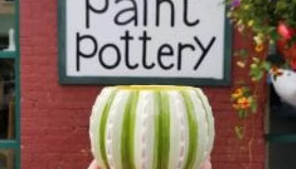 In Saratoga Springs, a person holds a green and white striped pottery bowl resembling a watermelon in front of a red brick wall. A Paint Pottery sign is visible in the background, alongside a hanging basket with colorful flowers.