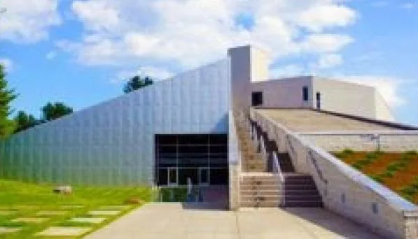 A modern building in Saratoga Springs boasts an angular glass facade and a sloped ramp on one side, set against a partly cloudy sky. The surrounding landscape is beautifully designed with lush green grass and trees.