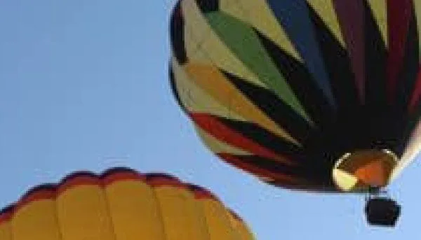 Two colorful hot air balloons gracefully float against the clear blue sky over Saratoga Springs. One balloon dazzles with vibrant stripes, while the other boasts a primarily yellow design adorned with a wavy red line.