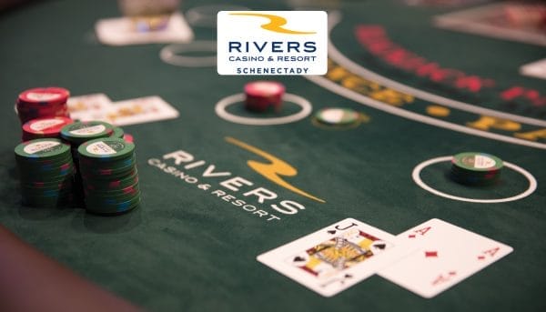 A poker table at Rivers Casino & Resort Schenectady, located in the heart of Capital-Saratoga, features poker chips stacked and two playing cards, a Jack of Spades and an Ace of Hearts, on the green felt surface. The casinos logo is visible in the background, inviting travelers to experience exciting tourism.