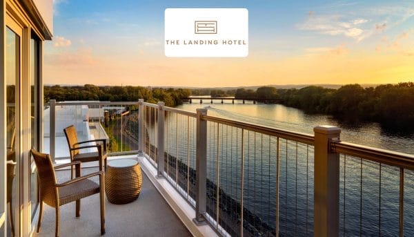 A hotel balcony overlooking a serene river at sunset in Saratoga Springs. The outdoor space features two wicker chairs and a small round table. The sky is filled with soft clouds, and the horizon showcases lush greenery. The Landing Hotel logo is at the top.