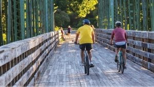 Two people wearing helmets ride bicycles across a wooden bridge on the Empire State Trail, surrounded by lush greenery. The sun shines brightly, casting long shadows. In the background, a few people stroll along the scenic path.
