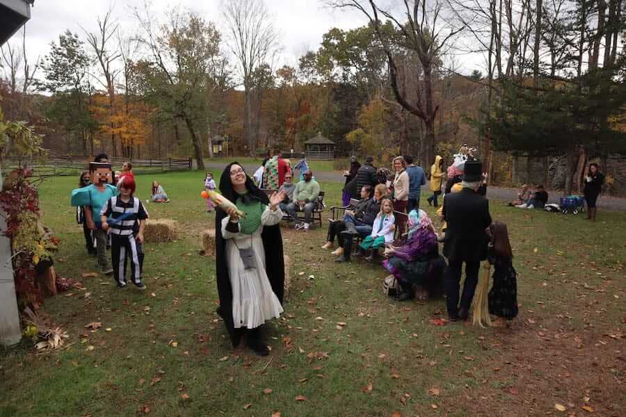A group of people enjoying Halloween at Ashokan gather outdoors in a grassy area adorned with hay bales. Some stand while others sit, surrounded by trees with autumn leaves under an overcast sky. Props and colorful decorations add to the festive spirit of the gathering.