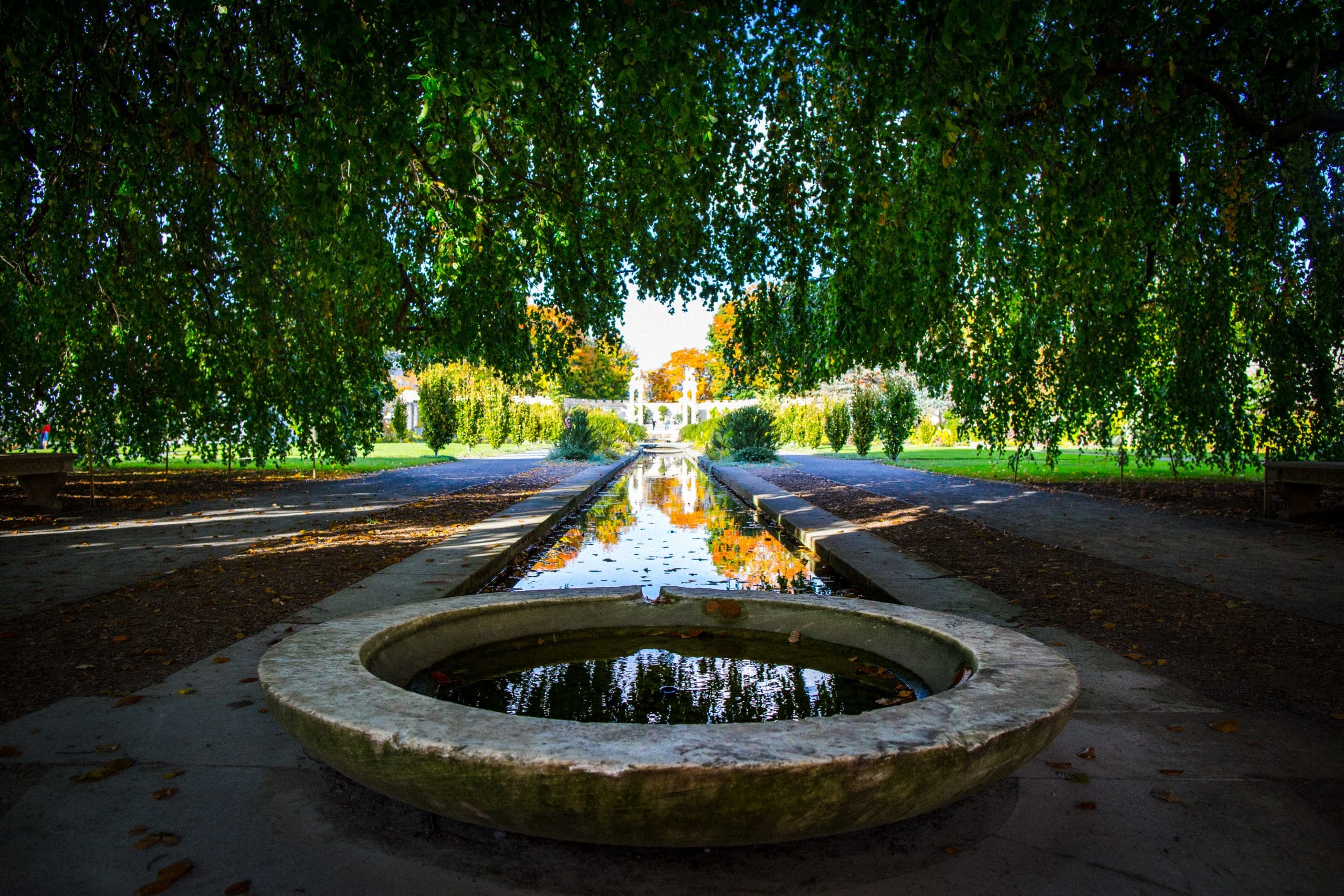 A circular stone fountain sits in the foreground at Untermyer Gardens Conservancy, reflecting overhanging green branches. Behind it, a narrow canal stretches through a lush garden, leading toward distant trees and a sunlit path.