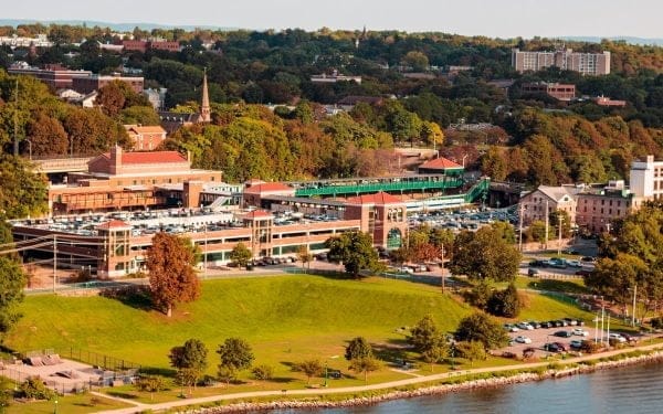 Aerial view of a cityscape featuring a large train station surrounded by trees and greenery. The station is adjacent to a parking area and near a body of water. Residential buildings and a church are visible in the background.
