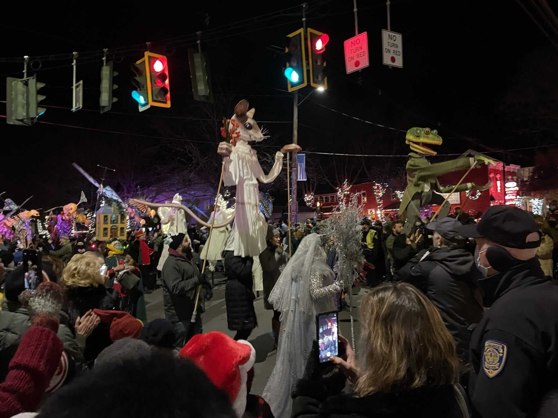A lively crowd gathers in Rhinebeck to watch a nighttime parade, where towering illuminated puppets and performers in festive costumes dazzle onlookers. Bright traffic lights shine above while holiday decorations light up the background, as people eagerly capture photos of the enchanting spectacle.
