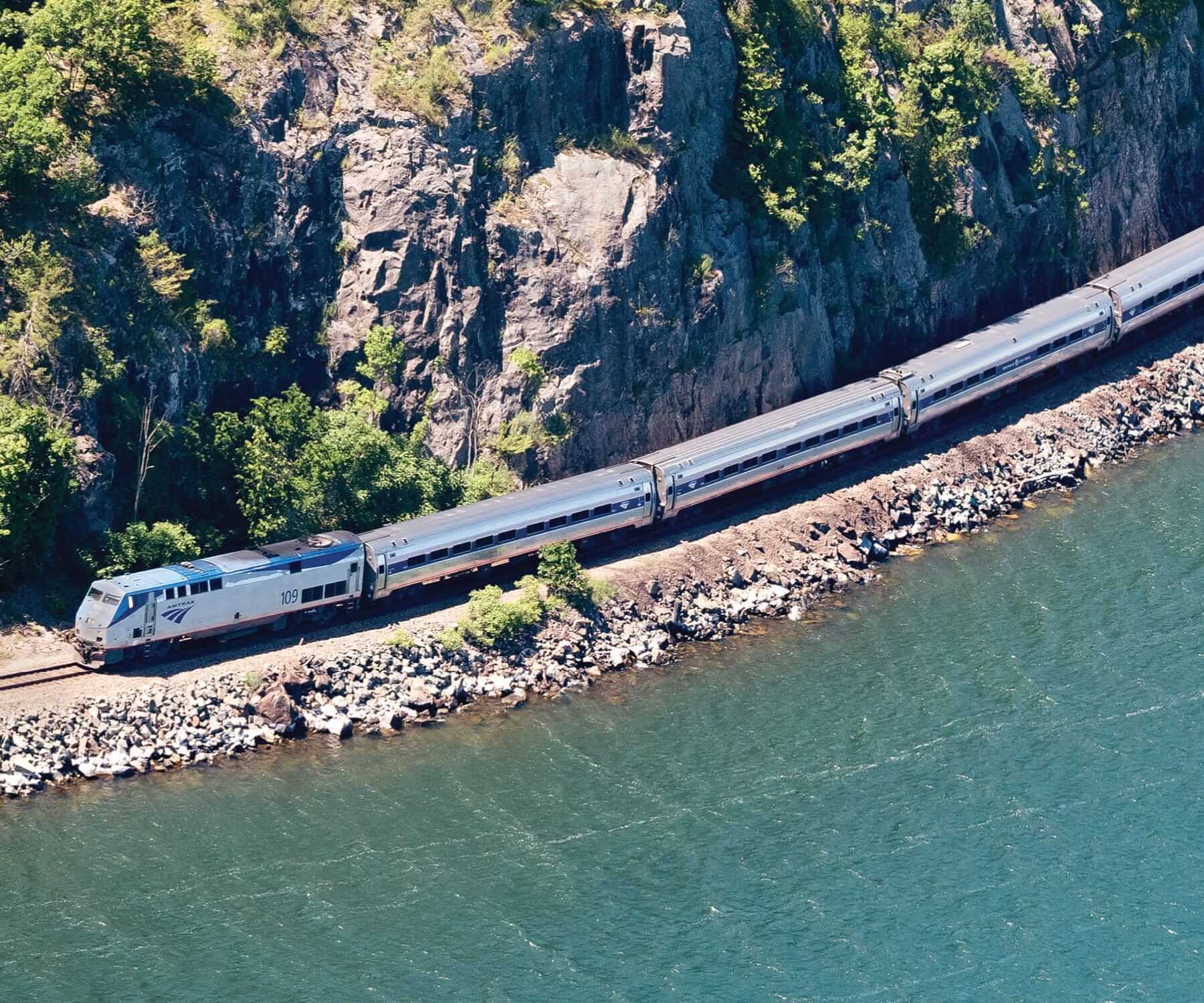 A passenger train on the Amtrak Adirondack Line travels along a rocky coastline, bordered by lush green trees and a body of water. The train curves around the landscape, following the railway tracks beside the shimmering blue water.