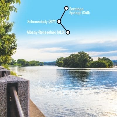 A calm river scene with trees lining the banks under a blue sky. A diagram showing connections between Saratoga Springs, Schenectady, and Albany-Rensselaer is overlaid on the sky.