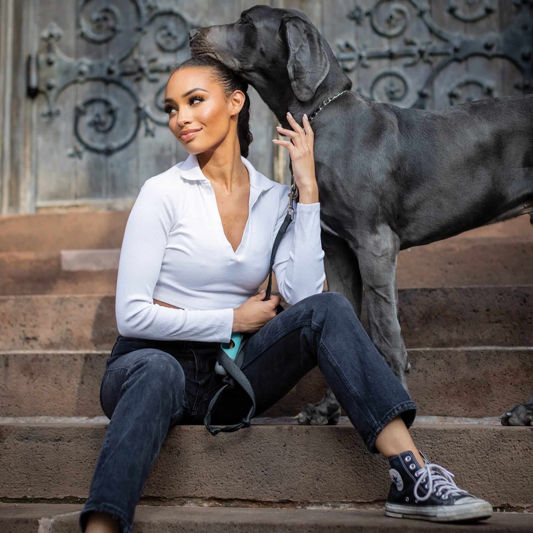 A woman embodies empowerment as she sits on stone steps in front of an ornate door, wearing a white shirt and dark jeans. She smiles warmly while a large grey dog, her loyal companion on this journey, leans affectionately against her head.