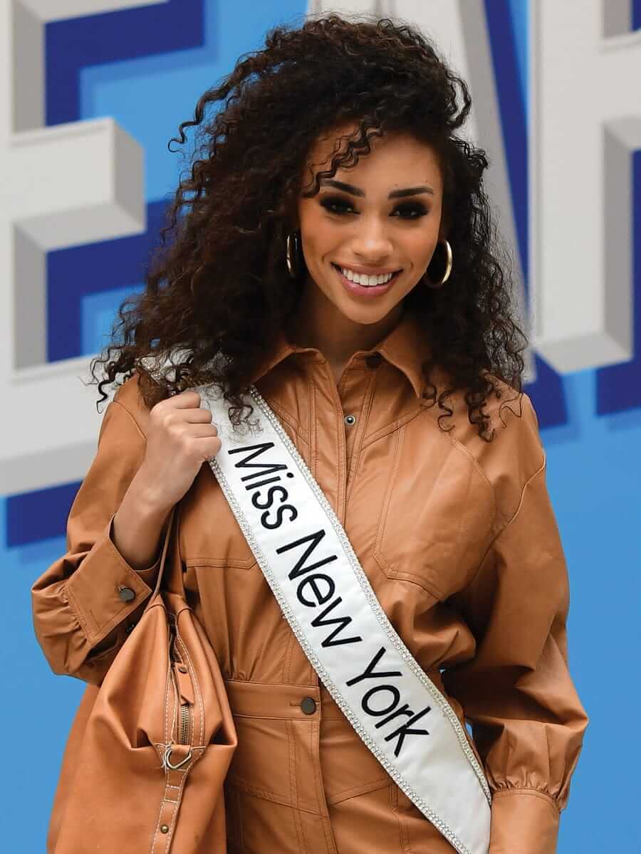 A woman with curly hair, wearing a brown outfit and a Miss New York Rising sash, smiles while standing in front of a blue and white background. She is holding a matching brown handbag, embodying her journey of empowerment.