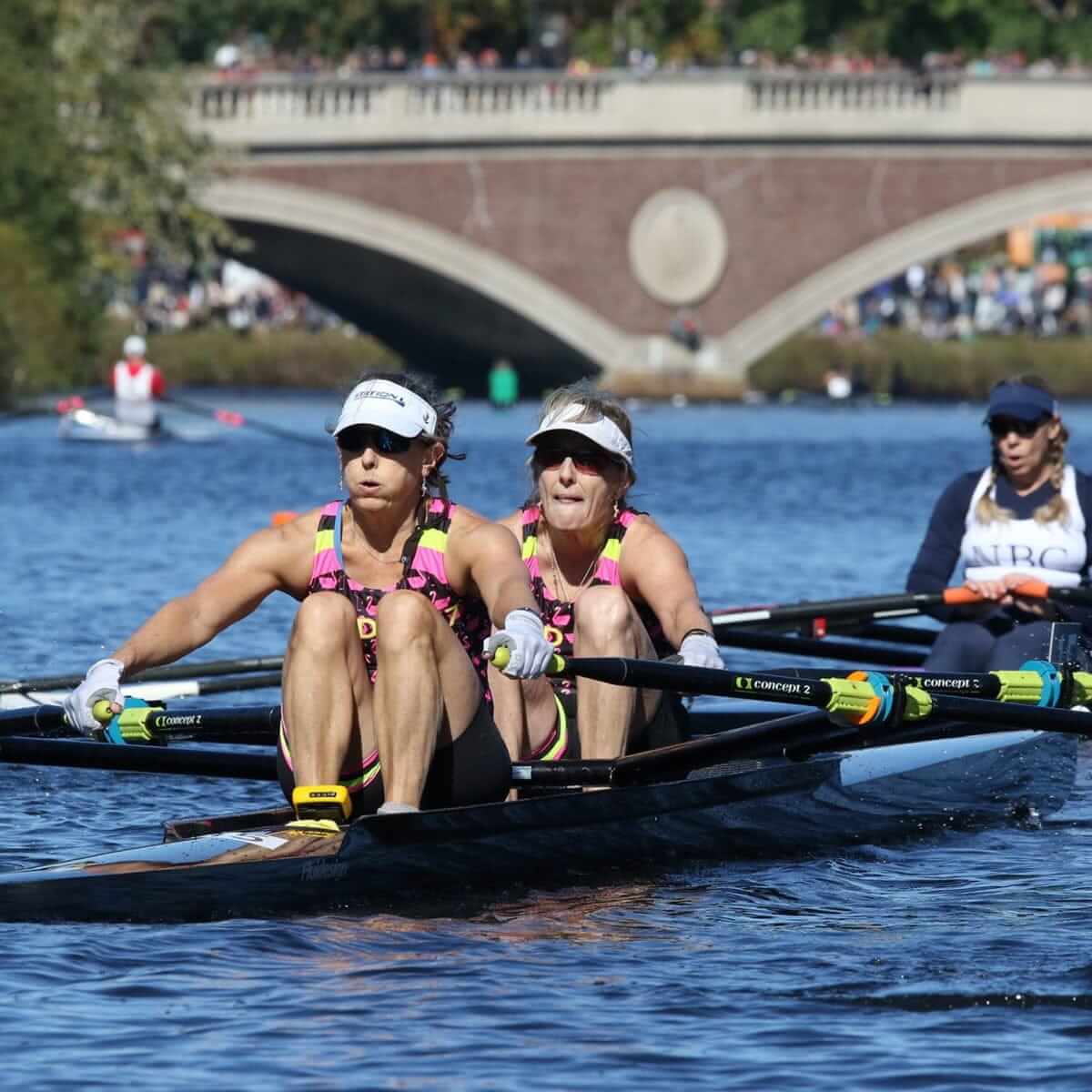 Two women rowers in a race on a river in Saratoga Springs, wearing matching gear and focused expressions, with a bridge and spectators in the background.