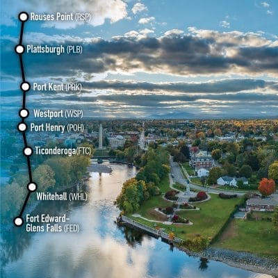 Aerial view of a scenic town with a river, autumn trees, and a partly cloudy sky. Text overlay shows a route map with stops at Rouses Point, Plattsburgh, Port Kent, Westport, Port Henry, Ticonderoga, Whitehall, and Fort Edward-Glens Falls.