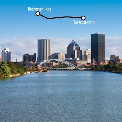 Skyline view of Rochester, NY, with tall buildings under a clear blue sky. A graphic line connects Rochester to Syracuse, with corresponding airport codes (ROC, SYR) labeled above. The Genesee River is in the foreground.