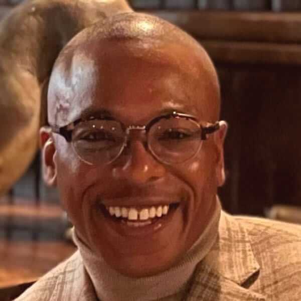 A person with glasses smiles warmly while wearing a plaid jacket, embodying the laid-back charm of New York City. The background is softly blurred, suggesting an indoor setting in the bustling Big Apple.