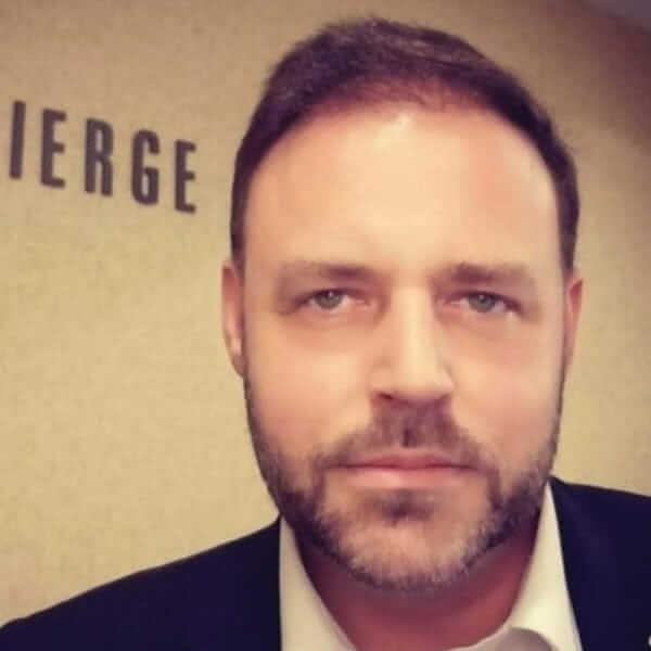 A man with a beard and short hair stands in a room with a beige wall, wearing a dark jacket over a white shirt. The partial word IERGE is visible behind him, subtly highlighting his recent trip to the Big Apple.
