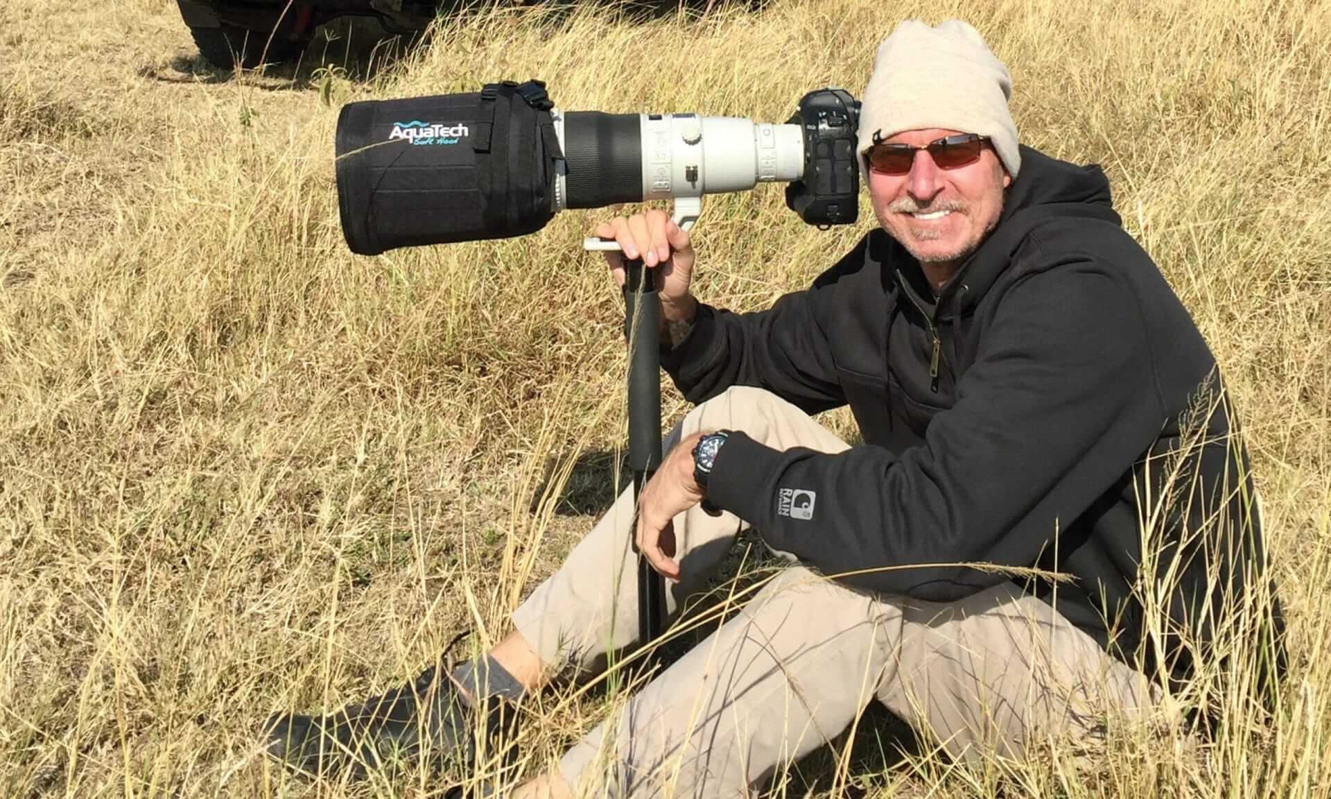 A person sits on dry grass, smiling and holding a large camera with a long lens, capturing that picture-perfect moment. They wear a beige hat, sunglasses, a black jacket, and light pants. The camera is securely mounted on a monopod, ensuring every shot hits the perfect pitch.