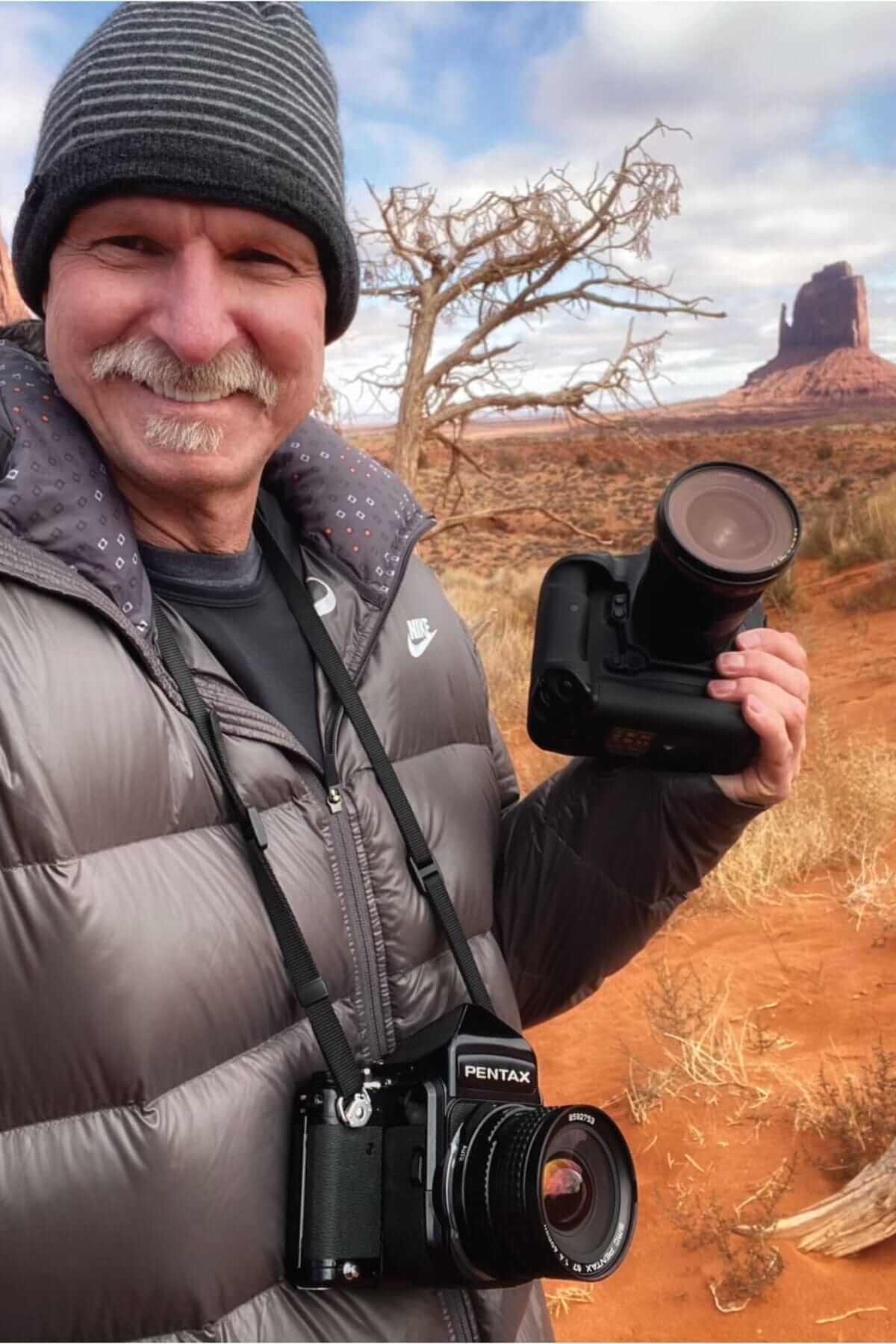 A person in a gray beanie and jacket smiles while holding a camera, capturing a picture-perfect moment. Another camera hangs around their neck as they stand in a desert landscape with reddish ground and a rock formation in the background. A person in a gray beanie and jacket smiles while holding a camera, capturing a picture-perfect moment. Another camera hangs around their neck as they stand in a desert landscape with reddish ground and a rock formation in the background.