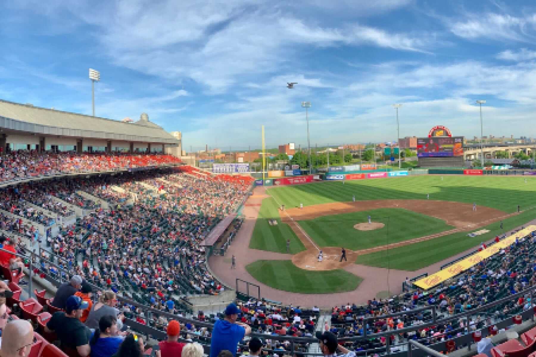 A bustling baseball stadium on a sunny day hosts a minor league game drawing a major impact with the spirited crowd. Players are on the field under blue skies with scattered clouds, and a large electronically lit scoreboard shines in the distance.