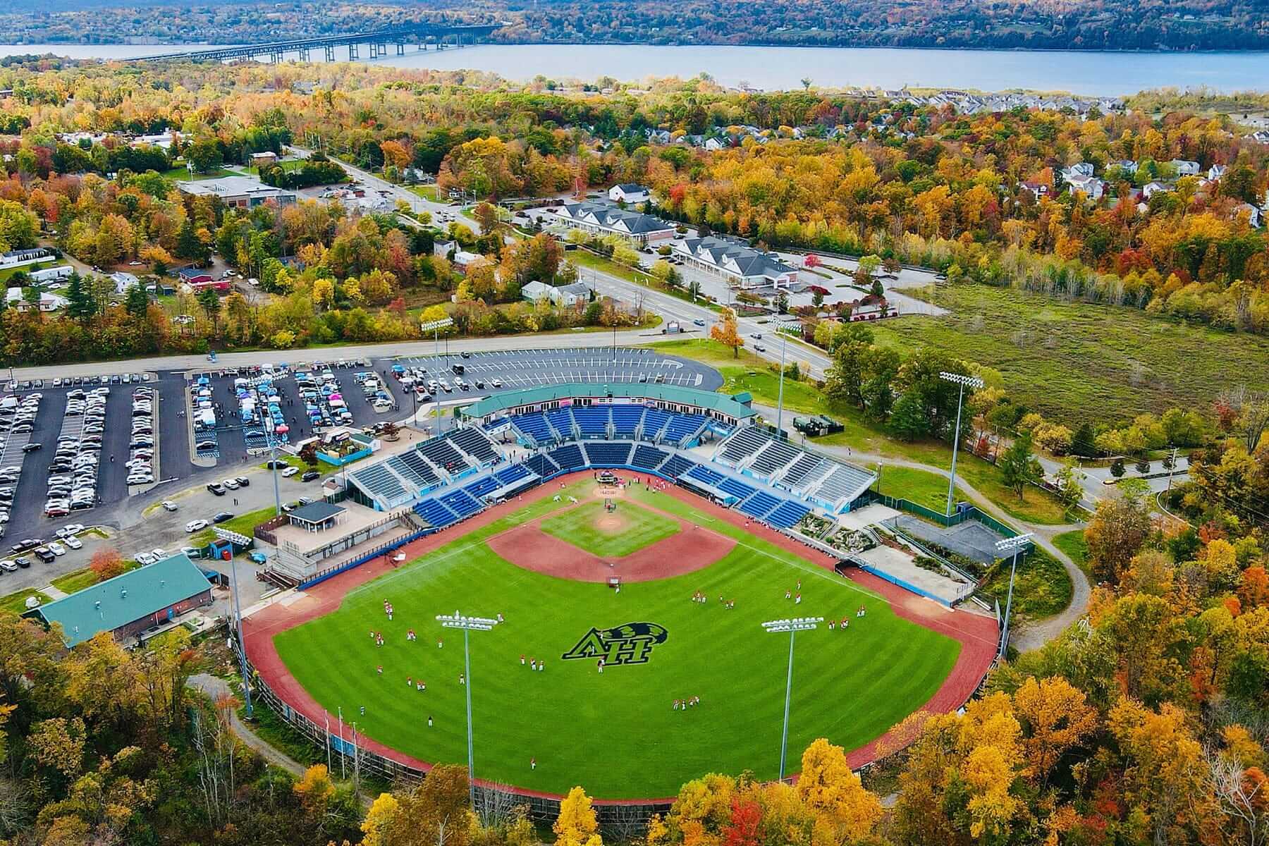 An aerial view of a Minor Leagues baseball stadium showcases a bright green pitch and blue seating areas, nestled among autumn-colored trees by a nearby river. Cars fill the adjacent lot, while houses and roads surround the area, highlighting this small venues major impact on the community.