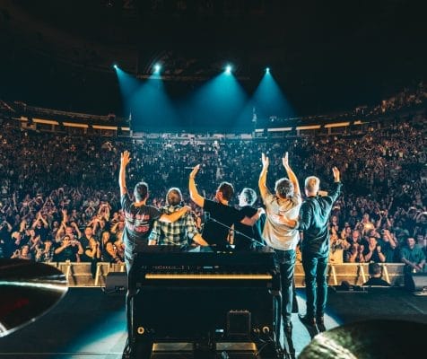 Amid a lively SPAC weekend, a band on stage waves to a cheering crowd in the large arena. Stage lights illuminate the scene, and a piano is visible in the foreground as the audience claps and raises their hands in excitement.