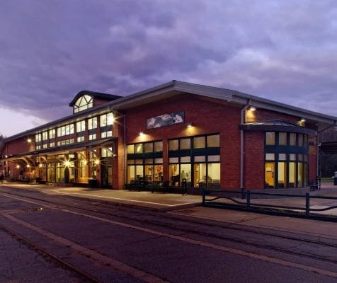 A brick building with large windows is illuminated at dusk, perfect for a quiet weekend reflection. The structure features a mix of traditional and modern design, with a gabled roof and outdoor lights lining the exterior. The cloudy sky creates a serene evening atmosphere.