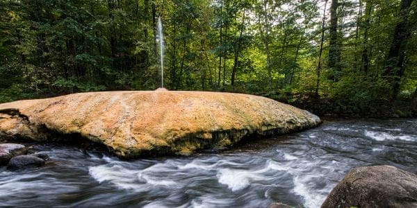 A natural mineral spring with a rocky, orange-brown mound is the perfect weekend retreat, surrounded by a flowing stream in a forest. The water spurts upward in a narrow jet, set against a backdrop of lush green trees and dappled sunlight.