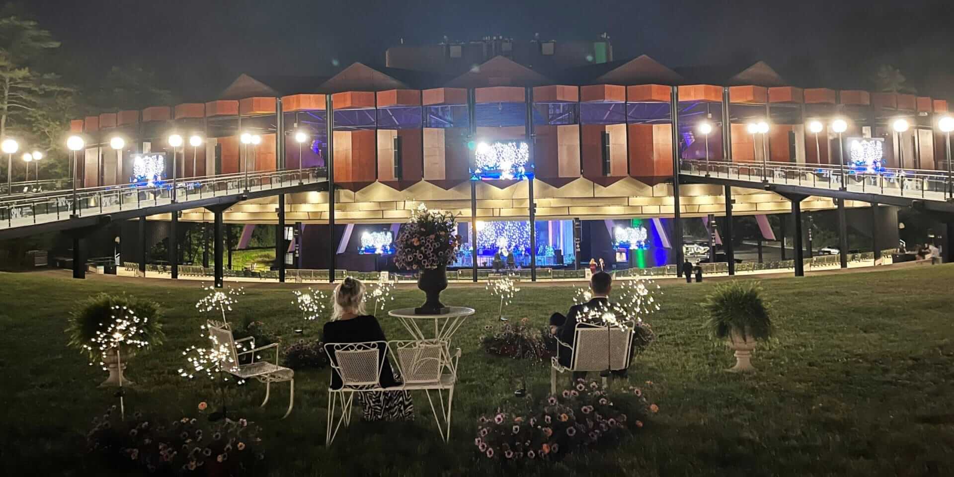 A nighttime scene showcases a large, illuminated building with unique architectural design and multiple balconies. In the foreground, people enjoy their weekend seated on outdoor chairs surrounded by decorative lights and plants, creating a vibrant SPAC-like ambiance.