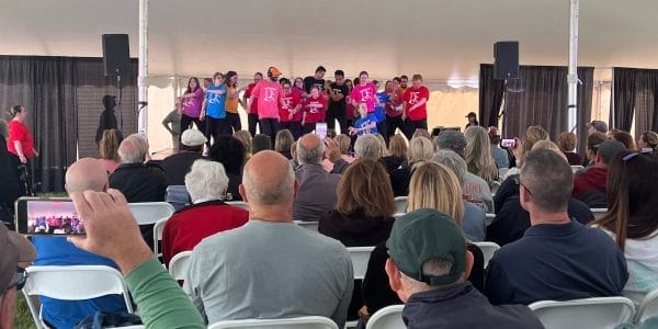 During the weekend, a group of performers in colorful shirts dazzles the audience under a large white tent at SPAC. Many spectators, seated and entranced, capture the vibrant display on their phones.
