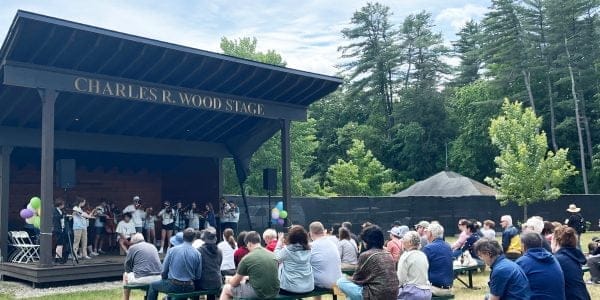 During the weekend, a group of musicians performs on the Charles R. Wood Stage at SPAC, playing string instruments. An audience seated on benches watches, set against a backdrop of trees under a partly cloudy sky. Balloons decorate the stage elegantly.