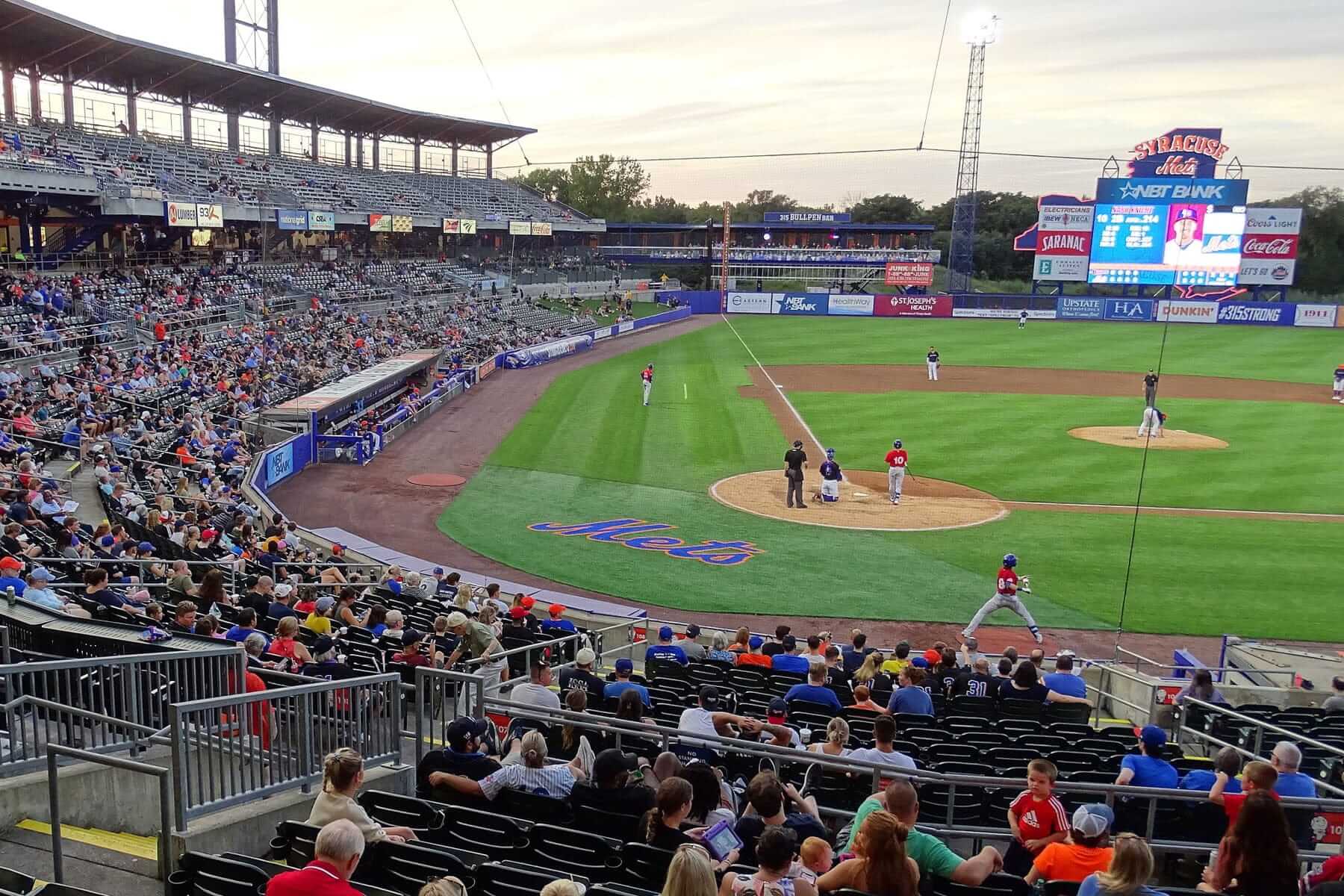 A lively baseball game is underway at a stadium with many spectators, showcasing the raw talent often seen in the Minor Leagues. The field shows players positioned at the bases and the batters box. The stands are filled with fans, and the scoreboard is visible in the background.