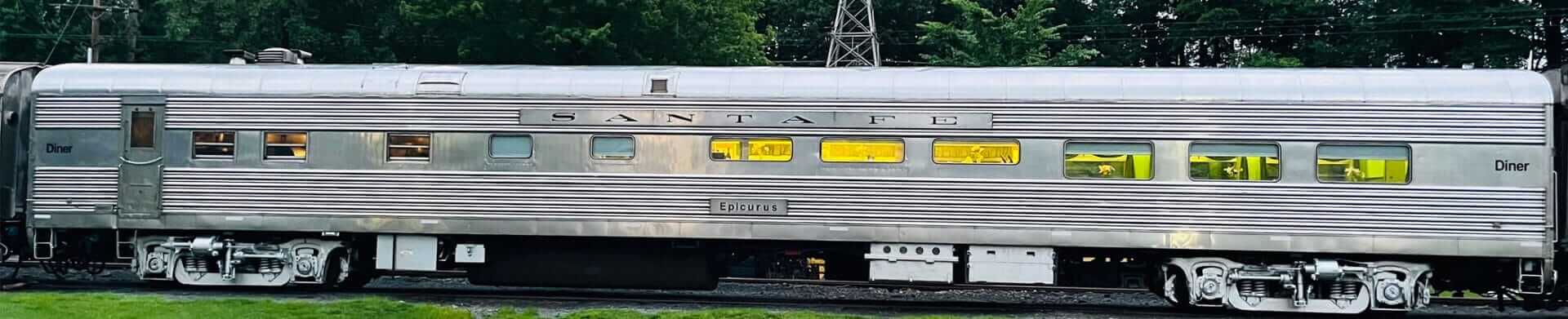 A vintage Santa Fe diner train car with a shiny silver exterior and lit interior stands stationary on the tracks, offering a taste of Saratoga Delights. Surrounded by lush green trees, it seems to be part of a classic railway exhibit, inviting onlookers to imagine a bygone era of train excursions.