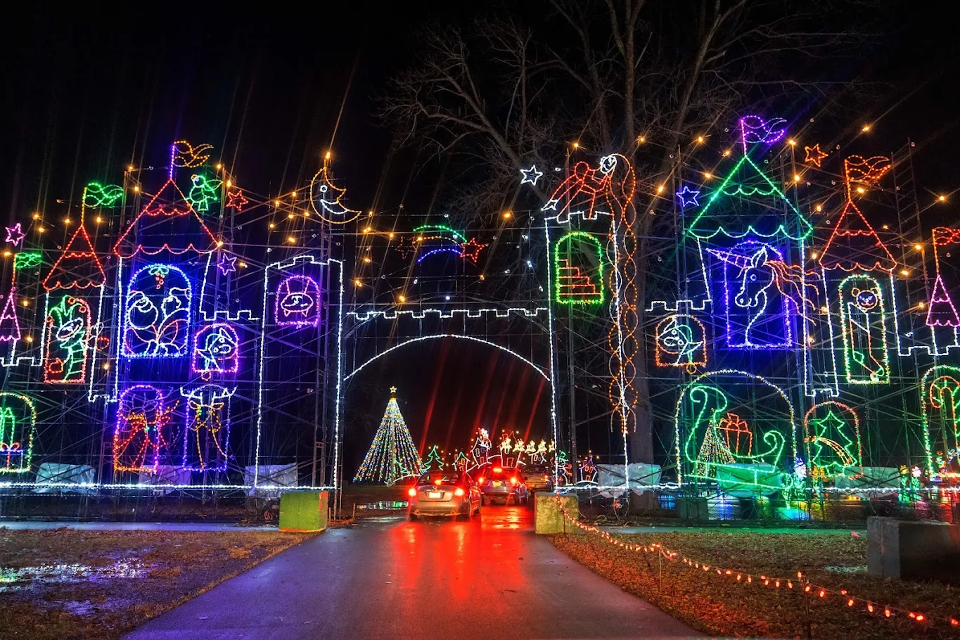 Cars drive through a brightly lit holiday light display shaped like a colorful castle at Onondaga Lake State Park, with arches, towers, and festive symbols. A decorated Christmas tree glows in the background.