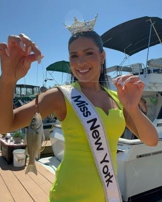 A woman in a bright green dress and a sash reading Miss New York smiles while holding a small fish on a line. She wears a crown and stands on a dock in the Great Northern Catskills, with boats gently bobbing under the clear blue sky, showcasing the charm of Greene County.