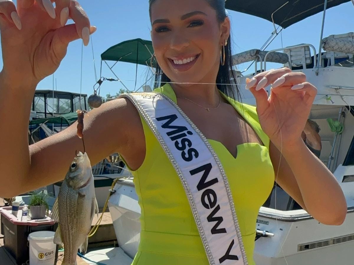 A woman wearing a Miss New York sash and crown stands smiling on a dock in the Great Northern Catskills, holding up a fishing line with a small fish. Boats bob gently in the Greene County background, creating a picturesque scene that captures her joy and love for nature.