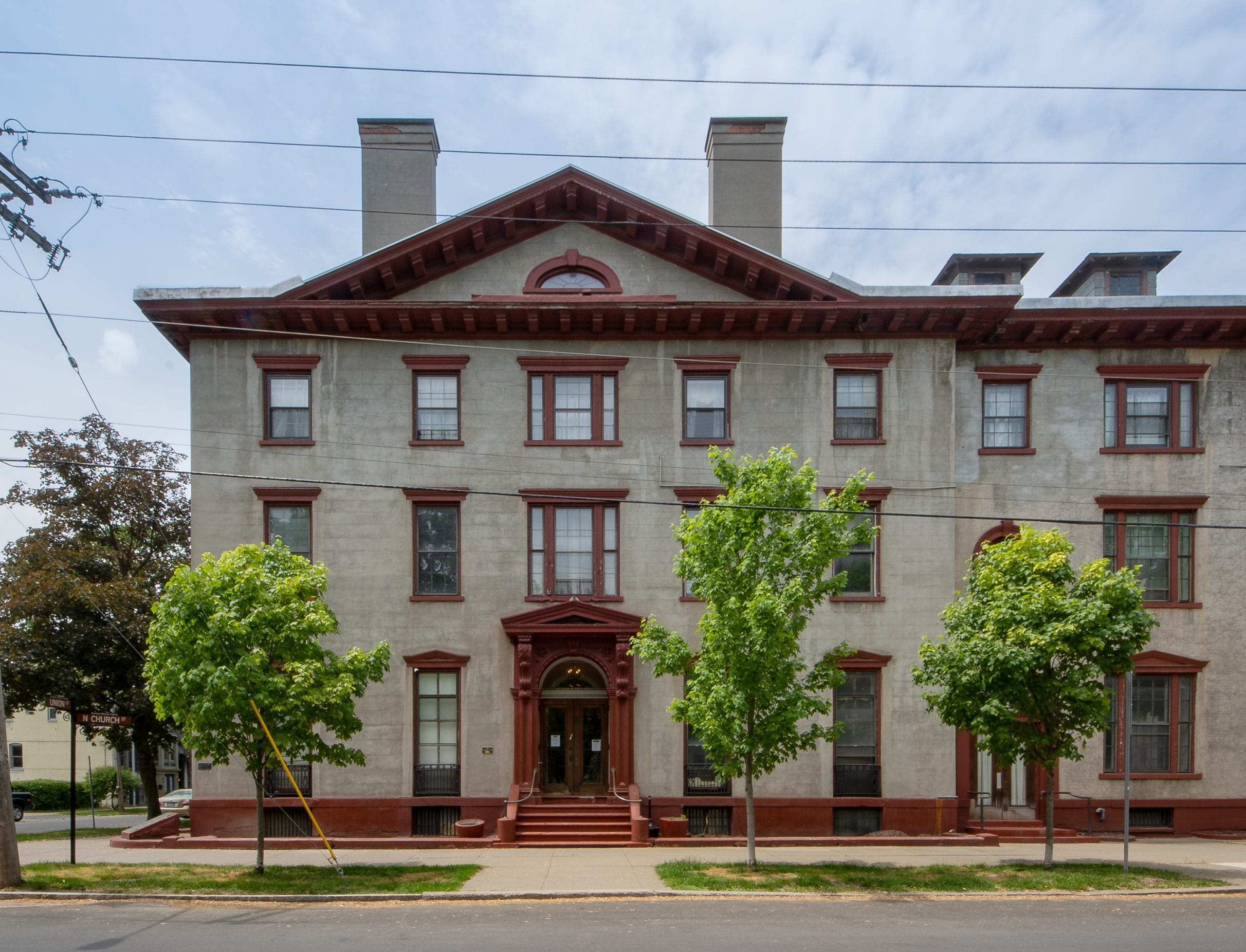 The historic three-story Stockade Inn features a gray facade, red trim, and large rectangular windows. Its symmetrical design includes a central arched entrance, with three leafy trees lining the sidewalk in front.