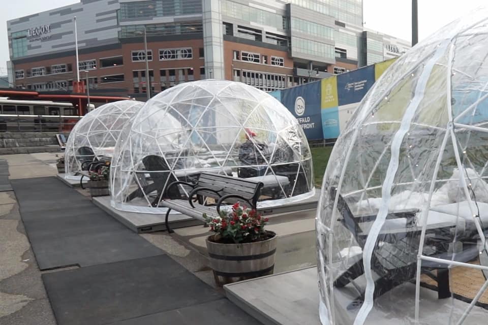 Clear plastic igloo domes with benches and tables inside are set up outdoors on a paved area in Buffalo NY, with potted plants nearby and modern office buildings in the background—perfect for a cozy New Year’s Eve Ball Drop celebration.