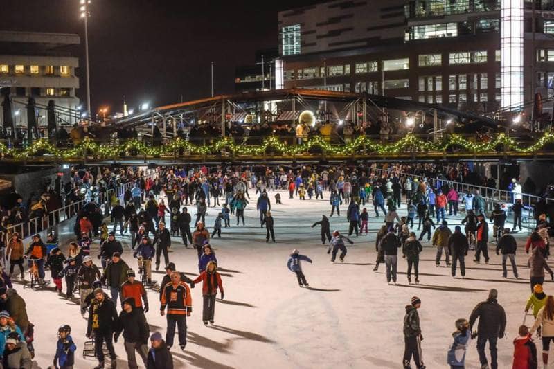 A large crowd of people ice skating outdoors at night under festive string lights and garland on a decorated bridge in Buffalo NY, with buildings lit up in the background.
